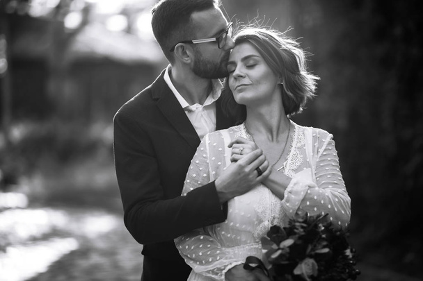 Beautiful black and white photo of bride and groom. Happy couple posing and smiling in wedding dress and suit with bridal bouquet in the park. People enjoy each other. Together. Wedding concept. - Фото, изображение