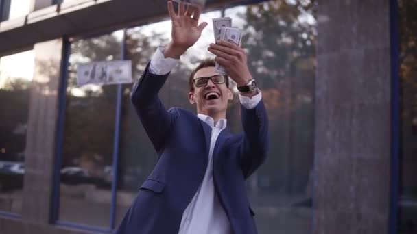 Caucasian businessman in blue suit throw handful of banknotes in the air. Super excited businessmen in glasses throw cash in front of office building and celebrate successful project and great deal - Materiaali, video