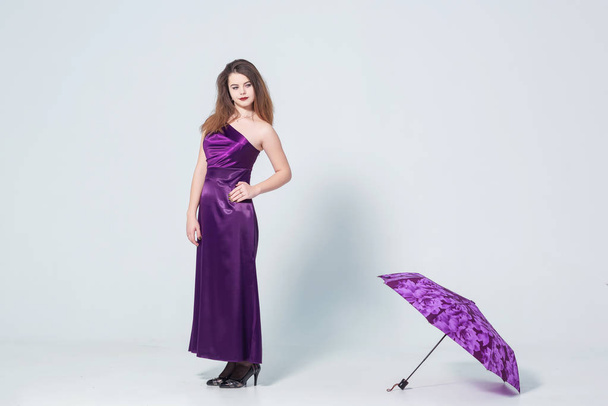 A girl in a purple dress on a white background with an umbrella in her hands. Girl and purple umbrella.  Young woman in evening dress on white background - Φωτογραφία, εικόνα