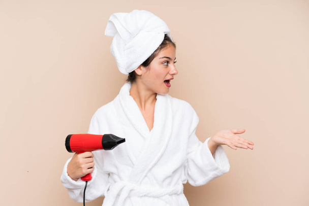 Young woman in a bathrobe with hairdryer over isolated background with surprise facial expression - Zdjęcie, obraz