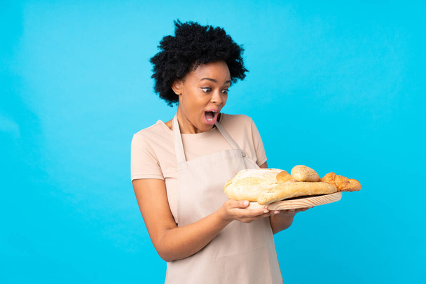 Afro-Amerikaanse vrouw in chef-kok uniform. Vrouwelijke bakker die een tafel met verschillende broden met verrassing en geschokte gezichtsuitdrukking vasthoudt - Foto, afbeelding