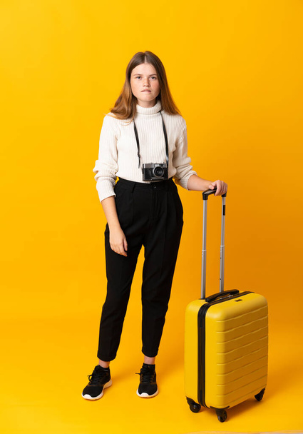 Full body of traveler teenager girl with suitcase over isolated yellow background having doubts and with confuse face expression - Foto, imagen