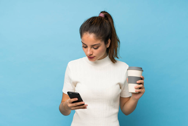 młoda brunetka dziewczyna na odizolowanym tle niebieski trzymając kawę do zabrania i telefon - Zdjęcie, obraz