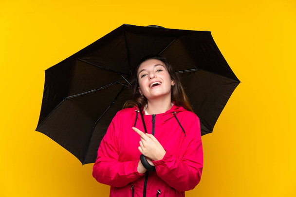 Joven morena sosteniendo un paraguas sobre un aislado fondo amarillo apuntando hacia un lado para presentar un producto
 - Foto, imagen