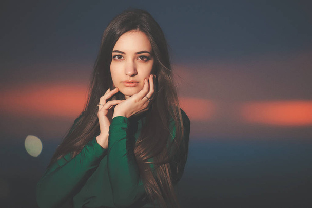 Portrait of beautiful young woman in long green dress on sunset background. Brunette girl at sunset. A girl with a beautiful face against the sky after sunset. - Valokuva, kuva