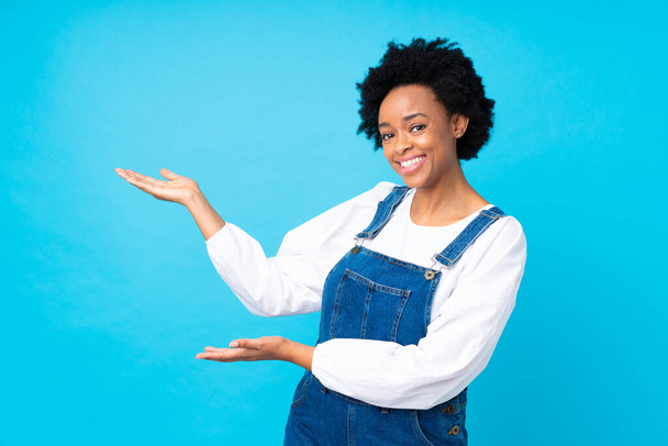 Femme afro-américaine avec des salopettes sur fond bleu isolé étendant les mains sur le côté pour inviter à venir
 - Photo, image