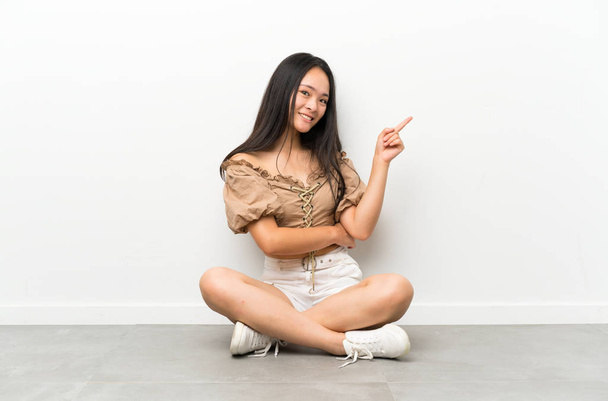 Подросток азиатская девушка сидит на полу указывая пальцем в сторону
 - Фото, изображение