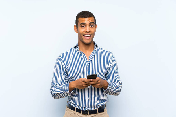 Νεαρός όμορφος μελαχρινός άντρας πάνω από απομονωμένο μπλε φόντο έκπληκτος και στέλνοντας ένα μήνυμα - Φωτογραφία, εικόνα