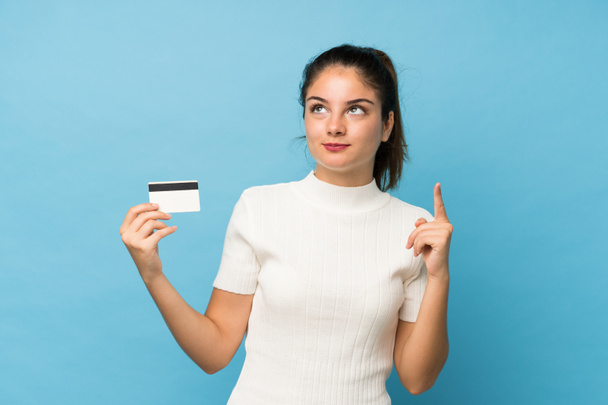 Mladá brunetka dívka přes izolované modré pozadí drží kreditní kartu - Fotografie, Obrázek