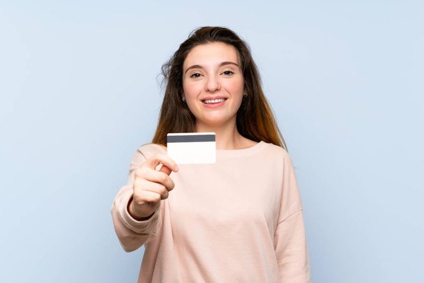 Young brunette girl over isolated blue background holding a credit card - Zdjęcie, obraz