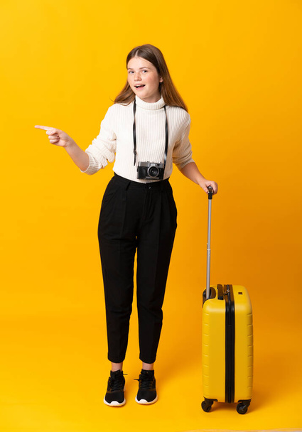 Koko kehon matkustava teini tyttö matkalaukku yli eristetty keltainen tausta yllättynyt ja osoittaa sormella sivulle
 - Valokuva, kuva