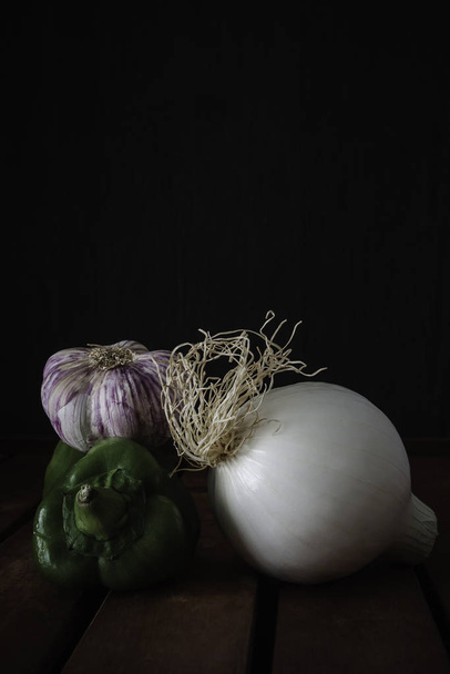 Zwiebeln, grüner Pfeffer und Knoblauch auf Holztisch und dunklem Hintergrund - Foto, Bild