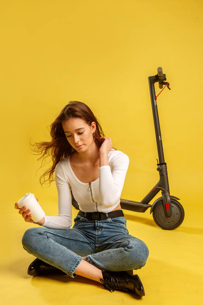 Young beautiful girl with electric scooter and coffee cup. Yellow background. Technology and freedom of movement. - Foto, Bild