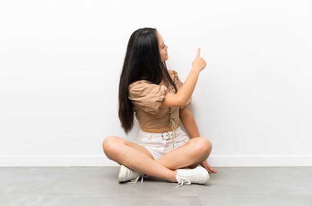 Adolescente ragazza asiatica seduta sul pavimento che punta indietro con l'indice
 - Foto, immagini