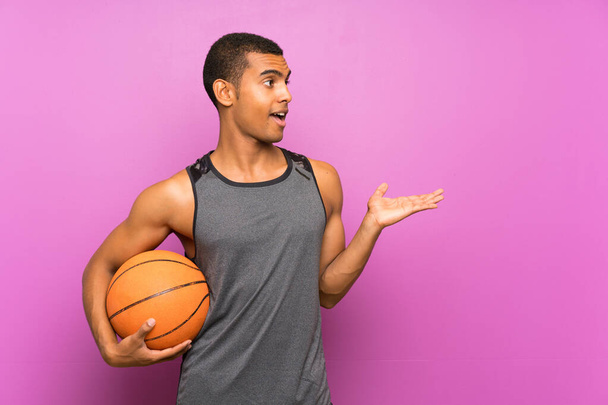Mladý sportovní muž s míčem basketbalu přes izolovanou fialovou stěnu s překvapivým výrazem obličeje - Fotografie, Obrázek