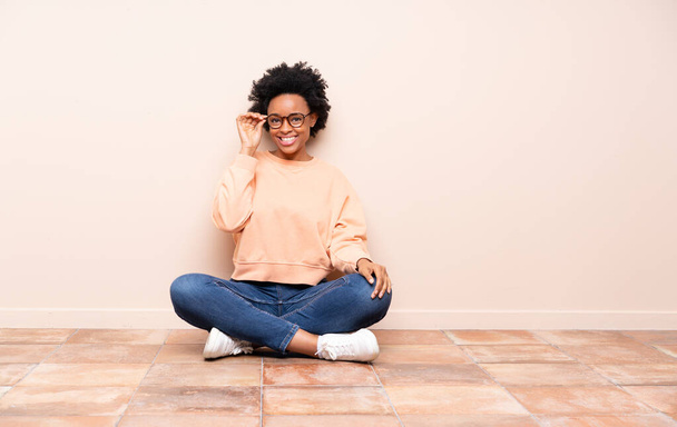 Αφροαμερικανή που κάθεται στο πάτωμα με γυαλιά και χαρούμενη. - Φωτογραφία, εικόνα