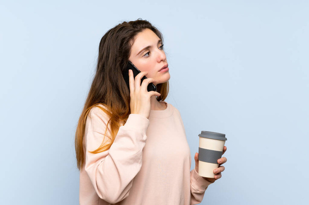 junge brünette Mädchen über isolierten blauen Hintergrund hält Kaffee zum Mitnehmen und ein Handy - Foto, Bild