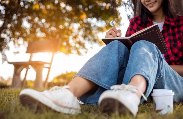 Молодая женщина пишет дневник, сидя под деревом в парке. Студент колледжа пишет записку
. - Фото, изображение