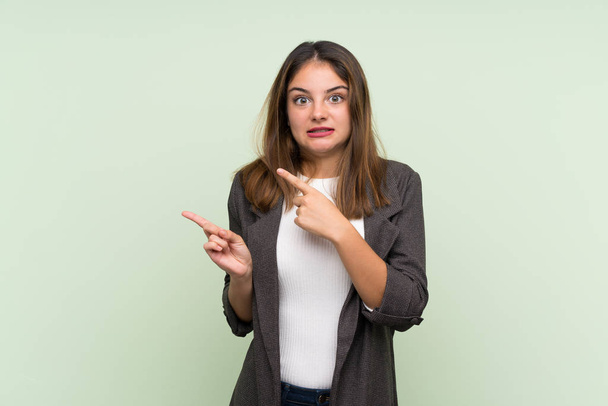 Young brunette girl with blazer over isolated green background frightened and pointing to the side - Photo, image