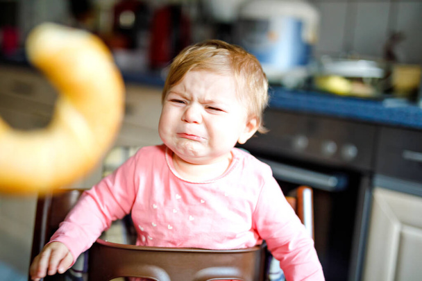 Aranyos kis bébi lány eszik kenyeret. Imádnivaló gyermek eszik az első alkalommal darab perec vagy croissant. Egészséges szomorú sírás gyermek. Első fogak jön. Otthon bent a konyhában. - Fotó, kép