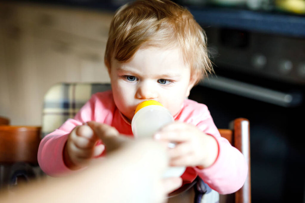Roztomilá roztomilá holčička držící kojící láhev a pijící mléko. První jídlo pro děti. Novorozené dítě, sedící v křesle domácí kuchyně. Zdravé děti a koncepce krmení láhví - Fotografie, Obrázek