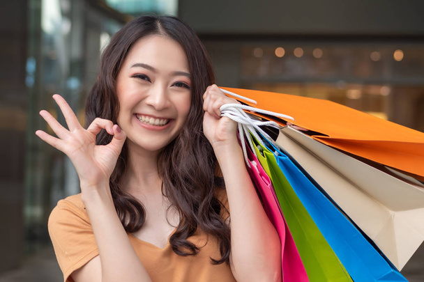 χαρούμενη χαμογελαστή γυναίκα ψώνια με πολύχρωμο τσάντα ψώνια στο εμπορικό κέντρο δείχνει ok σημάδι χέρι? έννοια της αγοράς, μεγάλη πώληση, ημέρα πυγμαχίας, πώληση γιορτή, έκπτωση, καλή συμφωνία για τις γυναίκες αγοραστές - Φωτογραφία, εικόνα