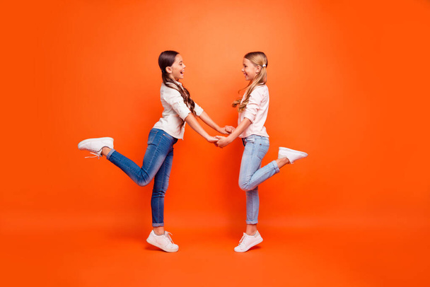 Full size profile side photo of positive cheerful two kids girls have free time hold hands rest relax wear modern clothing white sneakers isolated orange color background - Fotoğraf, Görsel