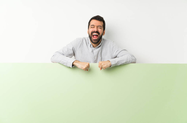 Joven hombre guapo con barba sosteniendo un gran cartel verde vacío gritando al frente con la boca abierta
 - Foto, Imagen