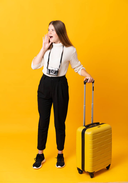 Full body of traveler teenager girl with suitcase over isolated yellow background shouting with mouth wide open - Foto, Bild