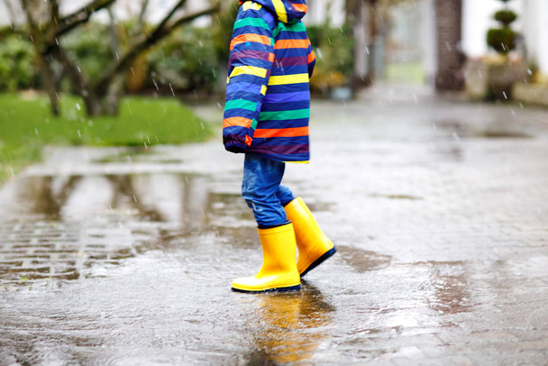 Sarı çizme giyen, yağmurda, yağmurda ve soğuk havada yürüyüş yapan çocuğun yakın çekimi. Renkli moda kıyafetleri içinde bir su birikintisine atlayan bir çocuk. Dışarıda eğleniyoruz. - Fotoğraf, Görsel
