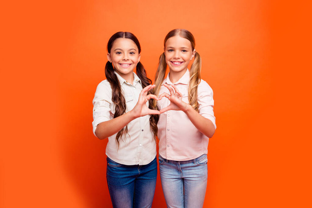 Portrait of positive cheerful two kids girl stand together make heart fingers sign of friendly loving family sister relationship wear casual clothing isolated orange color background - Valokuva, kuva