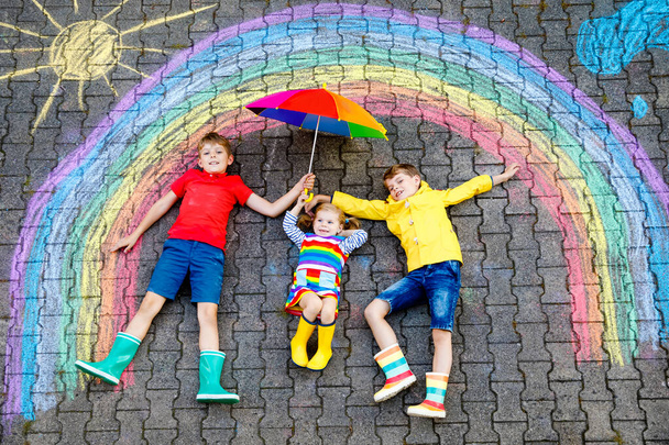 τρία μικρά παιδιά, δύο παιδιά σχολείο αγόρια και νήπιο κορίτσι που διασκεδάζουν με το ουράνιο τόξο εικόνα σχέδιο με πολύχρωμα κρητίδες στην άσφαλτο. Αδέλφια σε λαστιχένιες μπότες ζωγραφική στο έδαφος παίζοντας μαζί. - Φωτογραφία, εικόνα