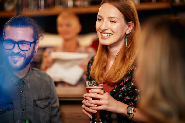 Portrait de gaie blonde caucasienne debout dans un pub, tenant une pinte de bière et bavardant avec des amis. Vie nocturne
. - Photo, image