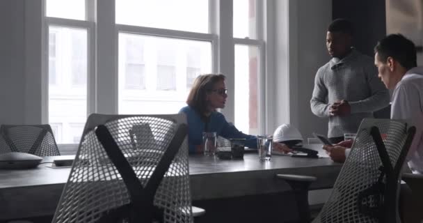 Vista lateral de una mujer de negocios caucásica sentada en un escritorio durante una reunión con un hombre de raza mixta sentado usando una tableta, y un hombre afroamericano de pie, mirando juntos los planos arquitectónicos y hablando en una oficina moderna
 - Metraje, vídeo