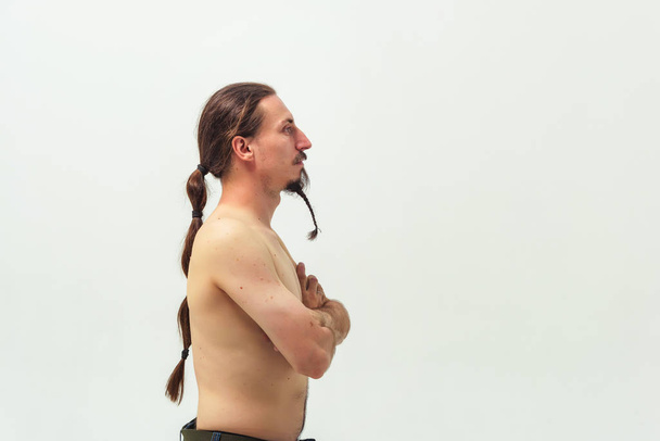 A man with long hair and a beard with a naked torso on white background side view in profile. Long hair gathered in a ponytail and a beard braid at the young man. - Φωτογραφία, εικόνα