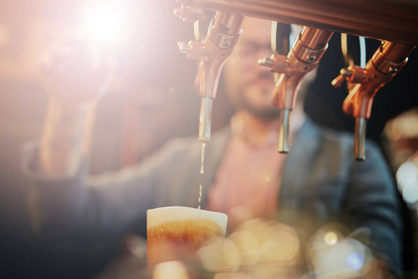 tätowierter kaukasischer Barmann, der Bier einschenkt, während er in der Kneipe steht. Selektiver Fokus auf Glas. - Foto, Bild
