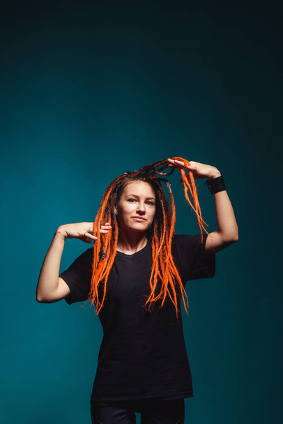 Girl with red fire dreadlocks on sea wave background color. A young woman with orange dreadlocks and piercings on a blue background. Beautiful daring informal image of a girl with red yellow dreadlocks - Fotó, kép