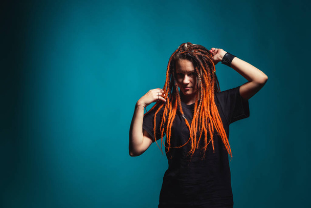 Girl with red fire dreadlocks on sea wave background color. A young woman with orange dreadlocks and piercings on a blue background. Beautiful daring informal image of a girl with red yellow dreadlocks - Photo, Image