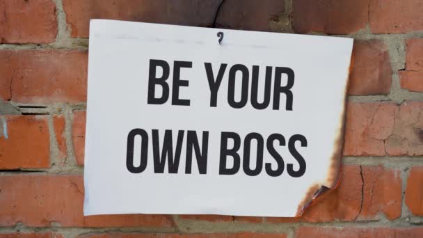 De inscriptie op een wit A4 formaat vel tegen de achtergrond van een oranje bakstenen muur. Het papier met het zwarte woord Be Your Own Boss brandt, rookt en verandert in as. Bedrijfsconcept. - Video
