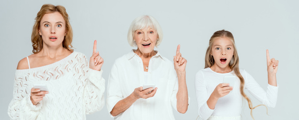 panoramic shot of shocked granddaughter, mother and grandmother holding smartphones and showing idea gestures isolated on grey  - 写真・画像