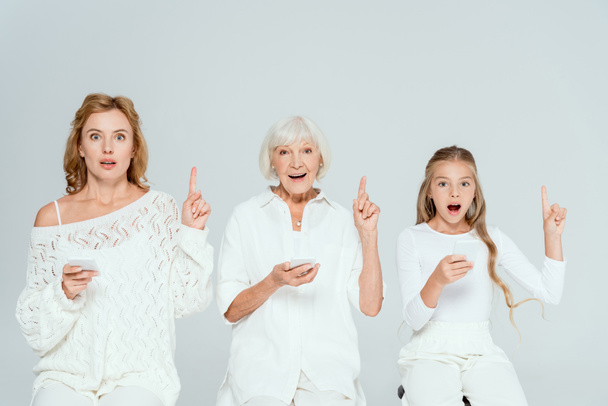 wstrząśnięta wnuczka, matka i babcia trzymające smartfony i pokazujące gesty idei odizolowane na szaro  - Zdjęcie, obraz
