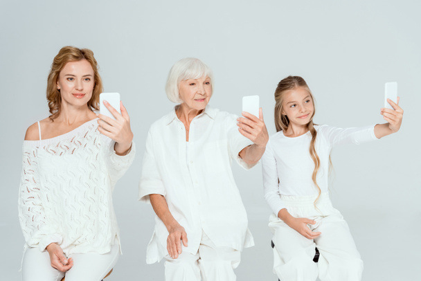 Lächelnde Enkelin, Mutter und Großmutter machen Selfies isoliert auf grau  - Foto, Bild