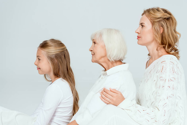 zijaanzicht van lachende kleindochter, moeder en grootmoeder knuffelen geïsoleerd op grijs  - Foto, afbeelding