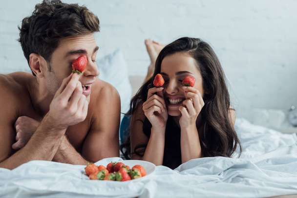 cheerful couple covering eyes with fresh strawberries while lying in bed - Фото, изображение