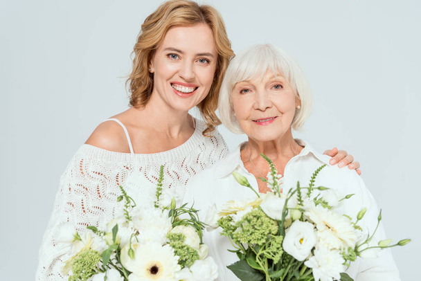 attractive mother and smiling daughter holding bouquets isolated on grey  - 写真・画像