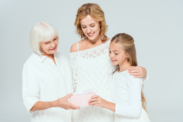 Lächelnde Enkelin, Mutter und Großmutter betrachten Geschenk isoliert auf grau  - Foto, Bild