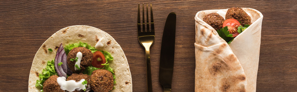 top view of falafel with vegetables and sauce wrapped and unwrapped in pita on wooden table with cutlery, panoramic shot - Photo, Image