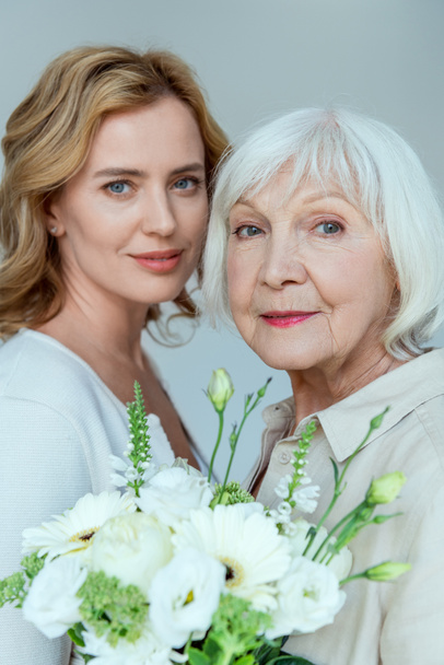 smiling daughter and mother with bouquet looking at camera isolated on grey  - Foto, imagen