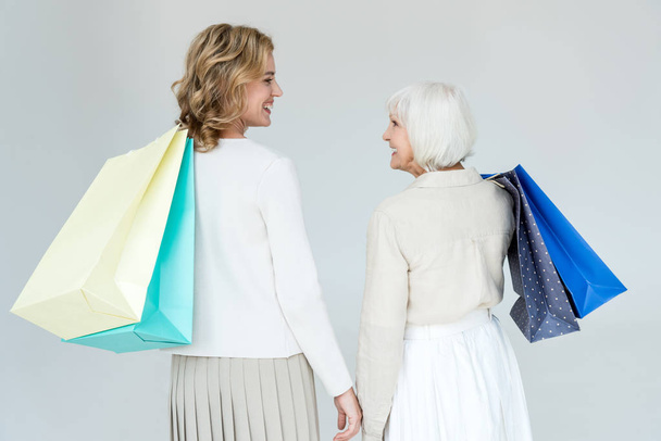 back view of smiling mother and daughter holding shopping bags isolated on grey  - Foto, imagen