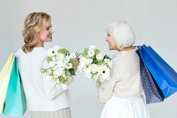 back view of smiling mother and daughter holding shopping bags and bouquets isolated on grey  - Fotografie, Obrázek